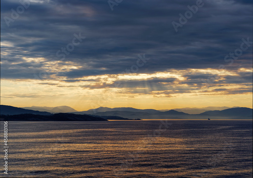 Rays of sunlight penetrating the low cloud layer above the Mountains and Islands of Bergen Fjord, with a Vessel in the Distance heading to Port.