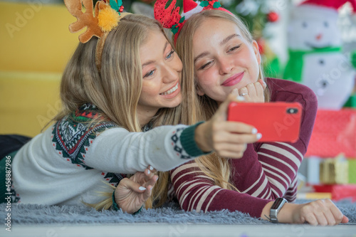 Two young caucasian girls with blonde hair wearing Christmas theme sweaters celebrating and enjoy Christmas at home while taking a selfie with red phone. Christmas trees in the background