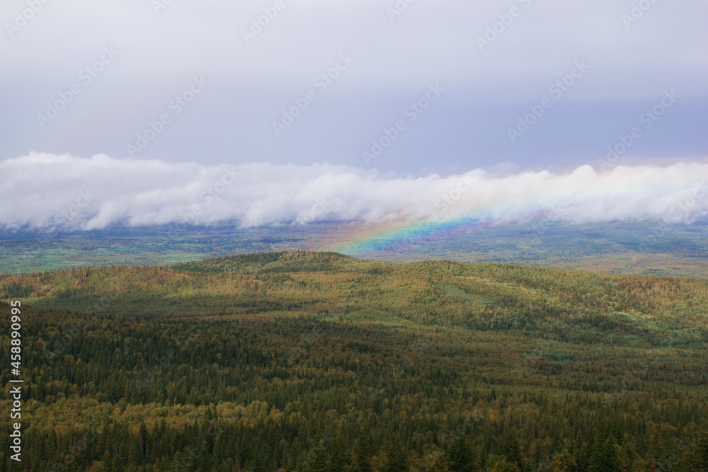 Scenic landscape with mountains and forest and cloudy sky with rainbow on background. Ural Mountains, Russia.
