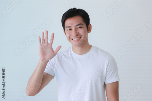 Studio portrait shot of Asian happy young handsome smart teenager teen guy model in casual clothing smiling show teeth look at camera waving hand greeting say hello hi of goodbye on white background photo