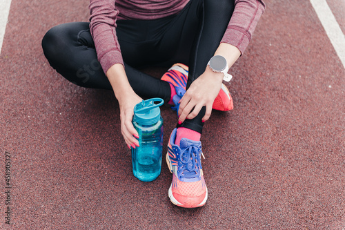 Sportswoman in stylish clothes and sneakers with bottle of water and smartwatch sits resting on track at stadium close view