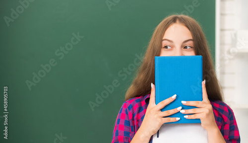 Photos teen girl in high school hiding behind workbook at blackboard, education