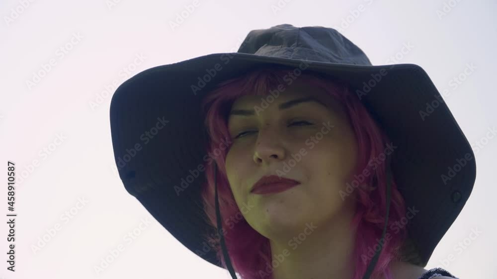 Mujer de pelo rosa con sombrero  grande y labios rojos, Mujer con sombrero mirando a los lados y sonriendo , chica joven con sombrero y de fondo el cielo blanco y nublado