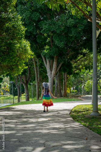 A little girl in a rainbow dress fun running on the concrete path in the park. View from behind.