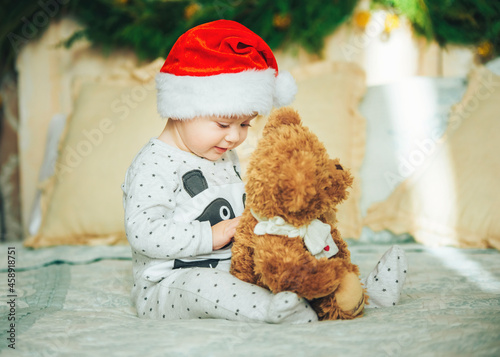 Funny baby boy wearing santa hat and pajama sitting on the bed with favorite toy. First Christmas. New Year's holidays