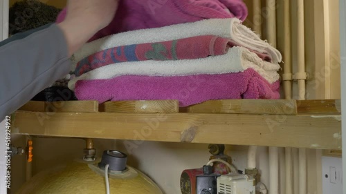 Closeup of the door to a domestic airing cupboard being opened, than a man's hand taking a clean towel from the pile on a slatted shelf above a hot water cylinder.