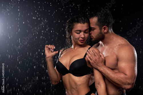 Portrait of passionate beautiful couple with athletic body under heavy rain on black background. sexy wet caucasian man kisses hugging woman while standing behind her. Love, passion concept © alfa27