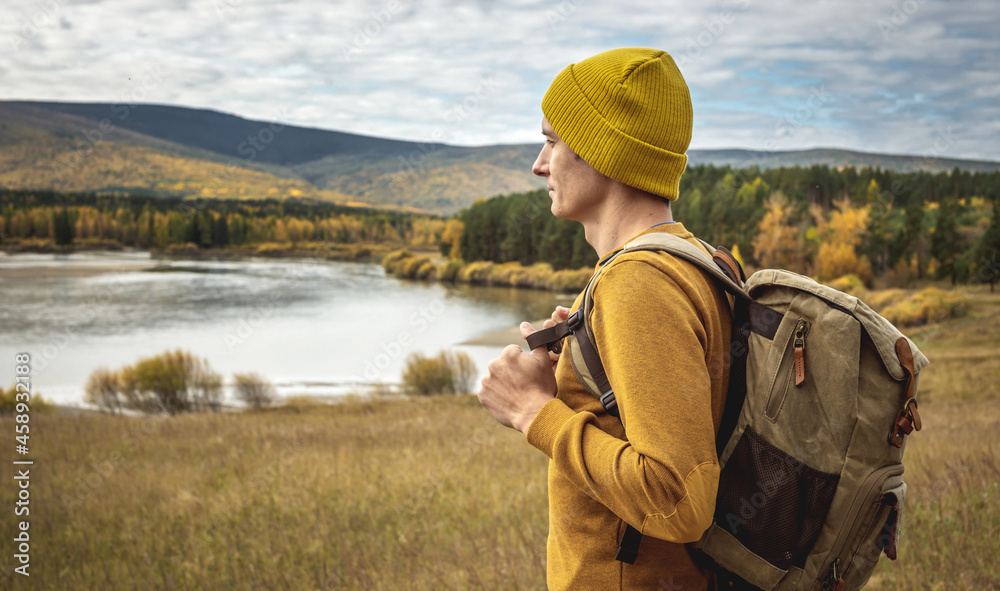 Man in a yellow hat and sweater with a backpack is standing in front of the river, autumn golden forest and hills Man in a yellow hat and sweater with a backpack is standing in front of the river, autumn golden forest and hills