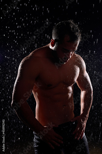 Stunning man with muscular body posing in rain, with naked torso, water drops on body. Young caucasian fit guy isolated on black background. Portrait. Sport, bodybuilding, fitness concept
