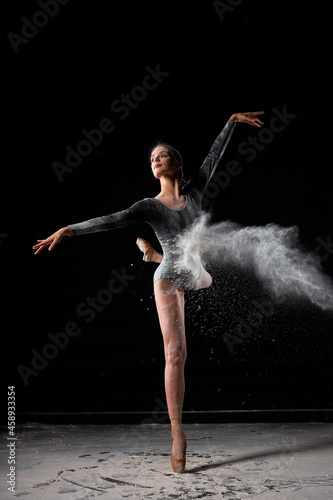 portrait of beautiful slender ballet dancer woman jumping, dressed in bodysuit, sensually moving among the flying flour which covers her body, on black studio background. Ballet, performance