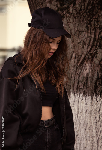Young thinking teen woman looking down and walking on the street tree background in black style clothing and cool cap. Autumn season clothing. Closeup © nastia1983