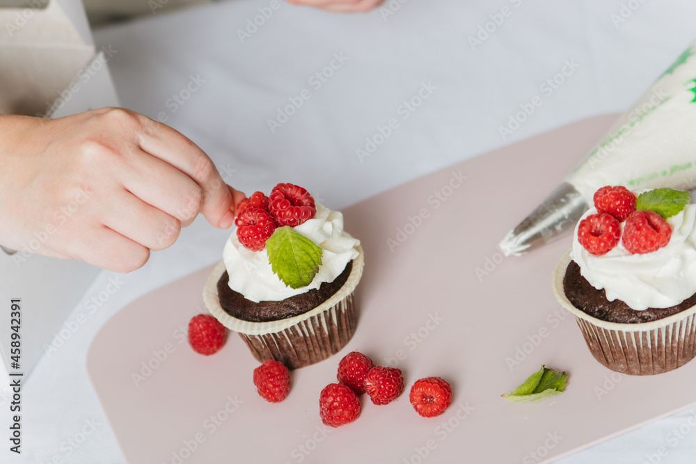 A woman prepares a cake with raspberries and white cream. Women's hands spread cream on a cupcake and decorate it with berries.