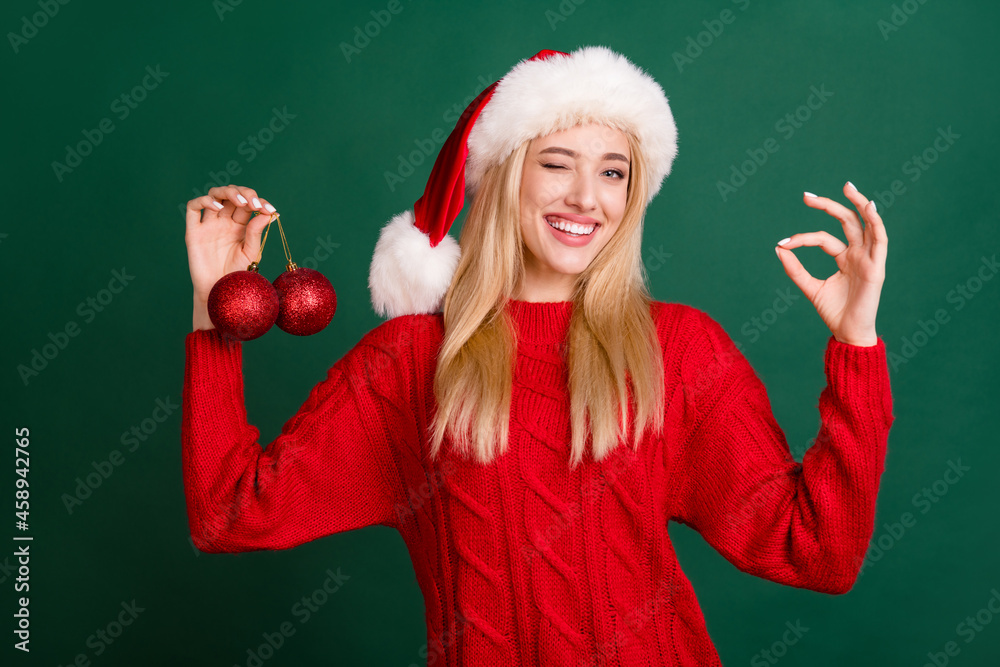 Photo portrait blonde woman in santa hat smiling hanging balls on xmas tree showing okay isolated green color background Photo portrait blonde woman in santa hat smiling hanging balls on xmas tree showing okay isolated green color background