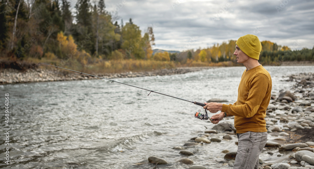 Man in a yellow sweater and hat is fishing on the river bank in the autumn forest with a spinning rod in his hands Man in a yellow sweater and hat is fishing on the river bank in the autumn forest with a spinning rod in his hands