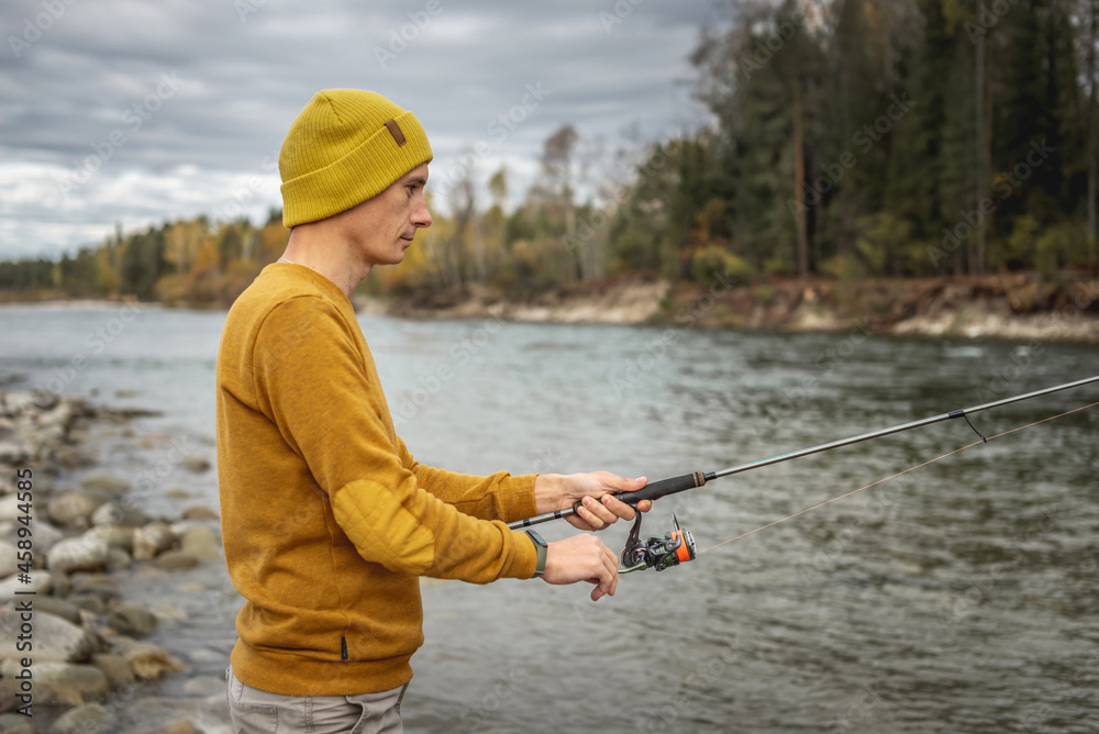 Man in a yellow sweater and hat is fishing on the river bank in the autumn forest with a spinning rod in his hands Man in a yellow sweater and hat is fishing on the river bank in the autumn forest with a spinning rod in his hands
