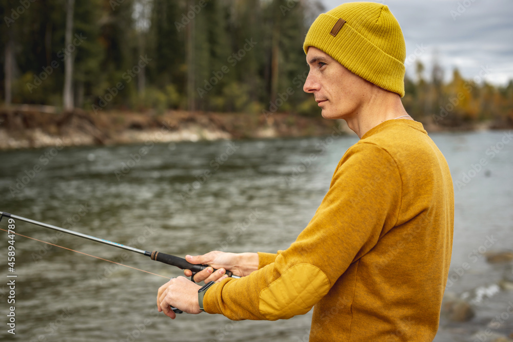 Man in a yellow sweater and hat is fishing on the river bank in the autumn forest with a spinning rod in his hands Man in a yellow sweater and hat is fishing on the river bank in the autumn forest with a spinning rod in his hands