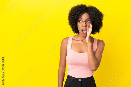 Young African American woman isolated on yellow background whispering something with surprise gesture while looking to the side