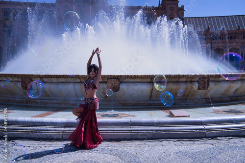 Middle-aged Hispanic woman, wearing a red dress with rhinestones, for belly dancing, dancing surrounded by soap bubbles in front of a water fountain. Belly dance concept.