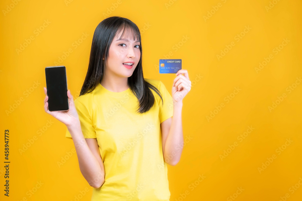 beautiful Asian young woman cute girl with bangs hair style in yellow shirt holding credit card and smartphone ,mobile phone uses a online banking for pay online shopping isolated on yellow background beautiful Asian young woman cute girl with bangs hair style in yellow shirt holding credit card and smartphone ,mobile phone uses a online banking for pay online shopping isolated on yellow background