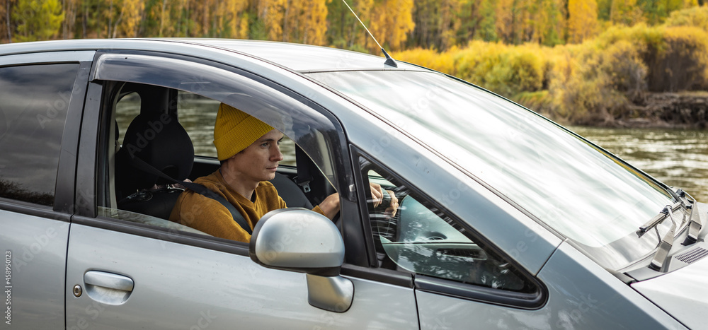 Obraz premium Man is driving a car along the river and the beautiful autumn forest. Concept of nature, travel and autumn mood