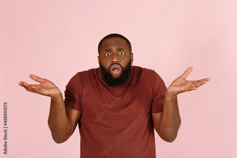 Confused young African-American person in brown t-shirt shrugs looking into camera on light pink background in studio closeup Confused young African-American person in brown t-shirt shrugs looking into camera on light pink background in studio closeup