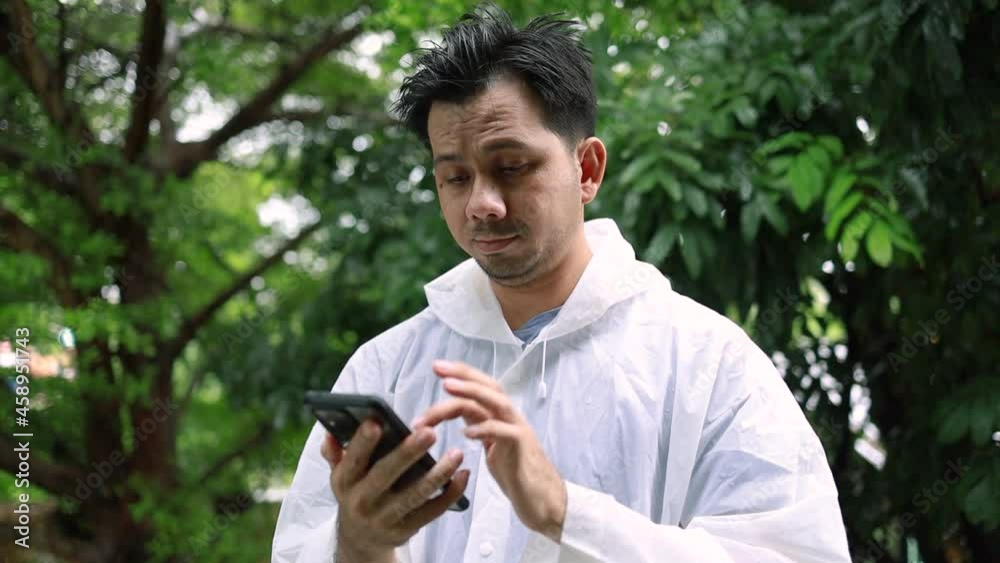 Asian man in a white raincoat standing using a smartphone outdoor Asian man in a white raincoat standing using a smartphone outdoor
