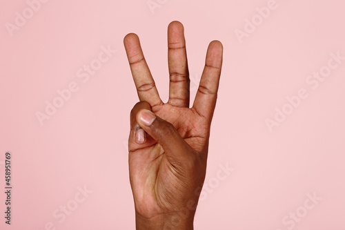 Young African-American man hand shows three fingers gesture on light pink background in studio extreme close view