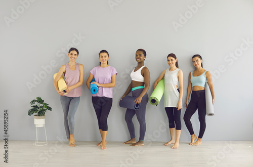 Sports and healthy. Portrait of happy different women standing in a row near the walls with yoga mats in their hands before playing sports in a bright gym. Concept of healthy lifestyle. Full length.