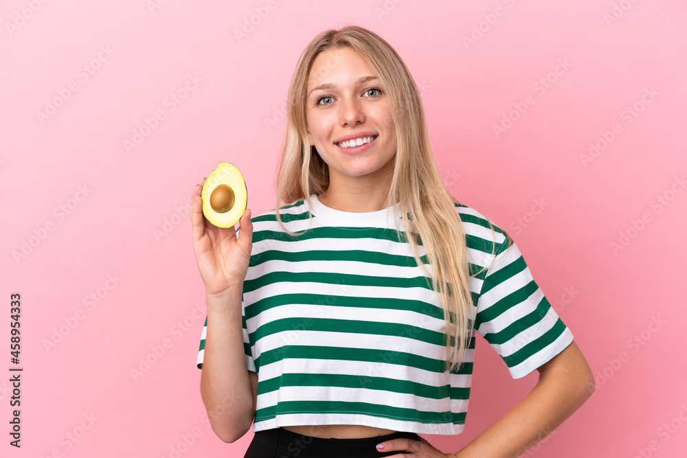 Young caucasian woman holding an avocado isolated on pink background posing with arms at hip and smiling Young caucasian woman holding an avocado isolated on pink background posing with arms at hip and smiling