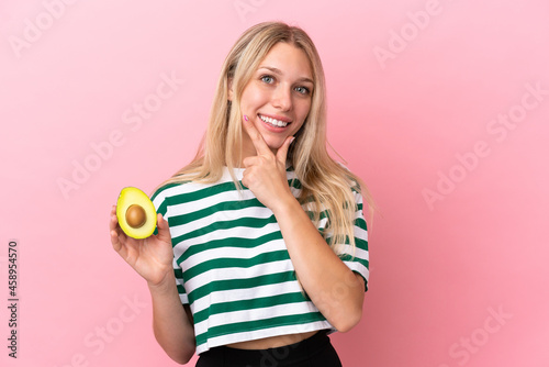 Young caucasian woman holding an avocado isolated on pink background happy and smiling