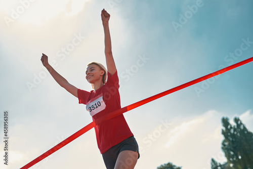 Low angle view of happy young female runner with arms raised reaching the finish line at track field during marathon outdoors