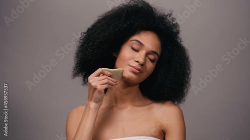 curly african american woman with bare shoulders using gua sha stone isolated on grey