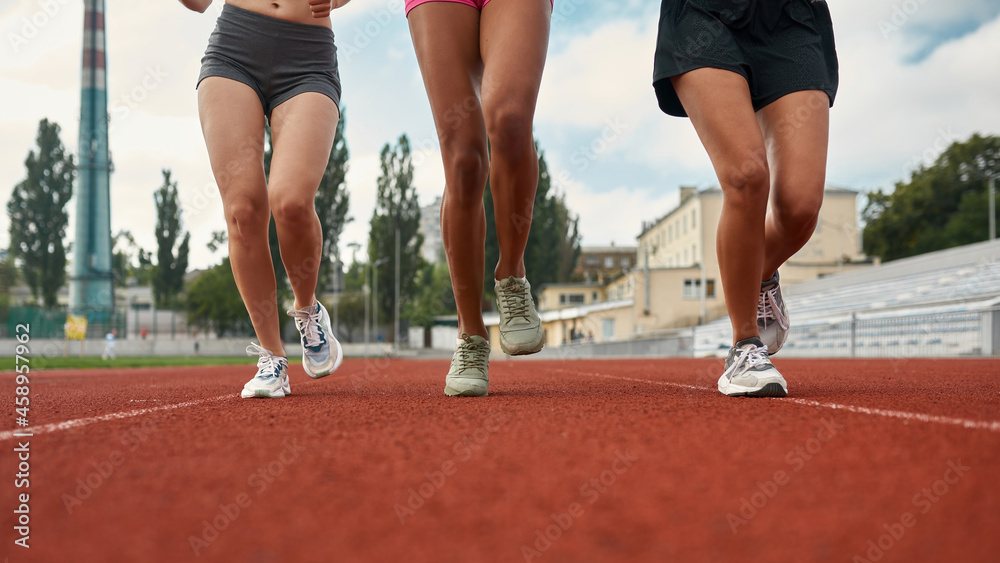Cropped shot of legs of three professional female runners in sportswear running together on track field at the stadium