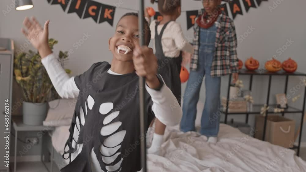 Slowmo tracking shot of 12-year-old African-American boy in Dracula costume and vampire teeth making spooky faces om camera while his friends jumping on bed during Halloween home party Slowmo tracking shot of 12-year-old African-American boy in Dracula costume and vampire teeth making spooky faces om camera while his friends jumping on bed during Halloween home party