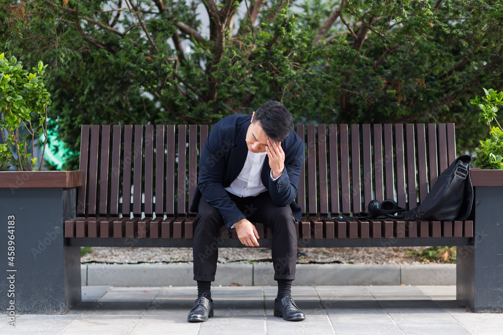 exhausted young asian business man office worker sitting on bench in city park outdoors. stressed overworked male with a headache massages head. Depressed Tired employe suffering pain sick work exhausted young asian business man office worker sitting on bench in city park outdoors. stressed overworked male with a headache massages head. Depressed Tired employe suffering pain sick work