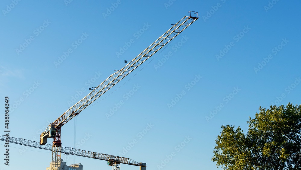 Construction site in the city centre with a couple of tall yellow cranes for lifting heavy construction material and tree on blue sky background. Construction industry. Space for text. Construction site in the city centre with a couple of tall yellow cranes for lifting heavy construction material and tree on blue sky background. Construction industry. Space for text.