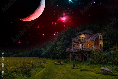 Nachtscène van een boomhut aan een bosrand en pad langs weide met warm geel lamplicht schijnt door de ramen tegen de achtergrond van de nachtelijke hemel met maan en helder twinkelende sterren photo