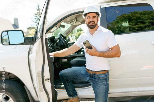 Man engineer builder wearing a white hard hat, shirt in front of his pickup using cellphone photo