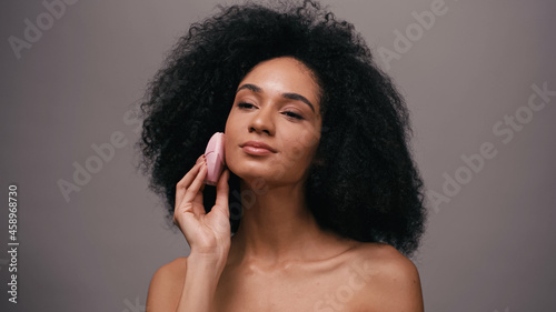 african american woman with bare shoulders using silicone cleansing device on face isolated on grey