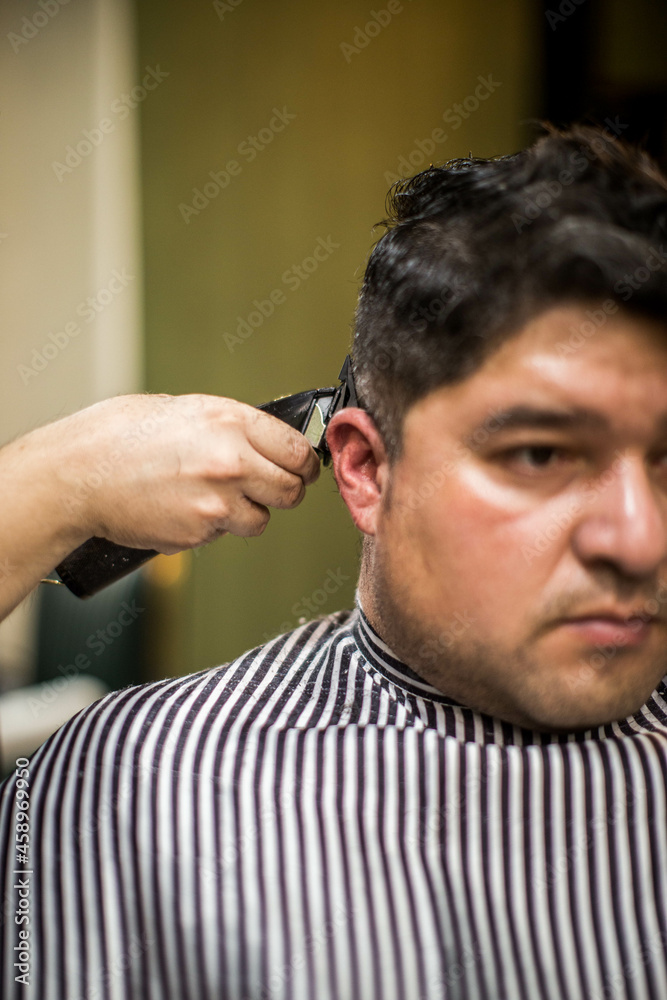 Obraz premium Man visiting hairstylist in barbershop. Barber works with hair clipper. Hipster client getting haircut. Hands of barber with hair clipper, close up