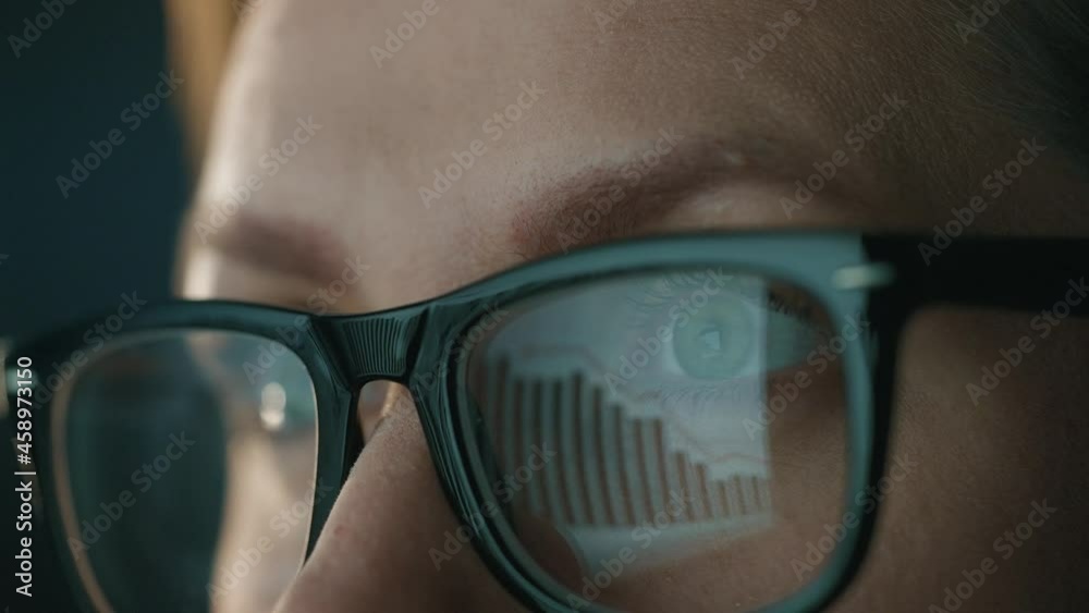 Woman in glasses looking on the monitor and and working with charts and analytics. The monitor screen is reflected in the glasses. Work at night. Extreme close-up