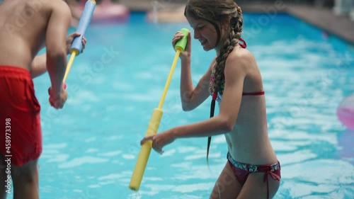 Carefree joyful Caucasian brother and sister playing with water gun standing in azure blue water in swimming pool. Happy cheerful children having fun with toys outdoors on sunny summer day