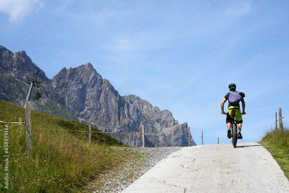 Obraz premium Man on a bicycle on a hilltop in rear view. There are Alp mountains on the left hand side. Cycling and hiking trail in region Engelberg in Switzerland.