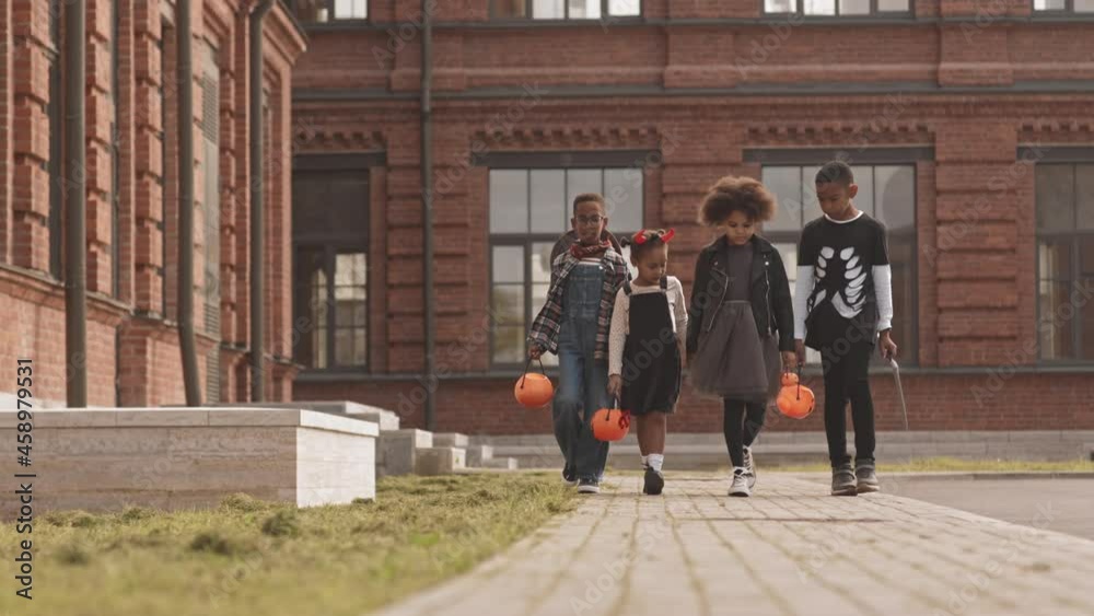 Slowmo shot of four cute African-American kids wearing Halloween costumes trick or treating outdoors running along street with pumpkin buckets holding hands Slowmo shot of four cute African-American kids wearing Halloween costumes trick or treating outdoors running along street with pumpkin buckets holding hands