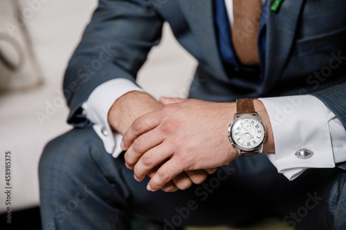 Grooms morning preparation Groom's in grey suit sitting with hand on hand Close up Confident groom holding hands together wearing a watch sitting on a chair resting his arms white background
