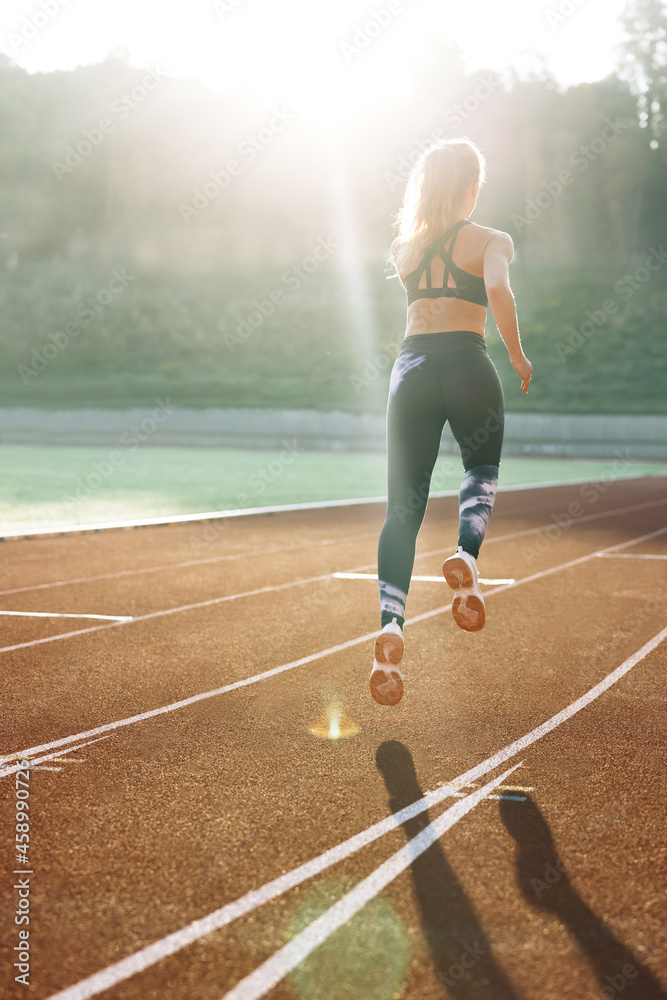 Obraz premium Rear view of athletic woman running on track stadium at summer morning light. Fit woman running on racetrack during training session. Female runner practicing on athletics race track. Sportswoman