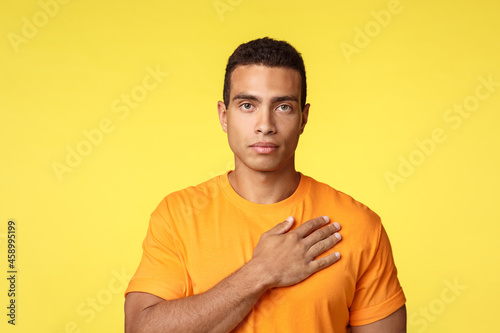 Young serious-looking and motivated sportsman hold hand on heart and standing as hear national anthem, show pledge or devotion gesture, look camera, keep memories in heart, yellow background