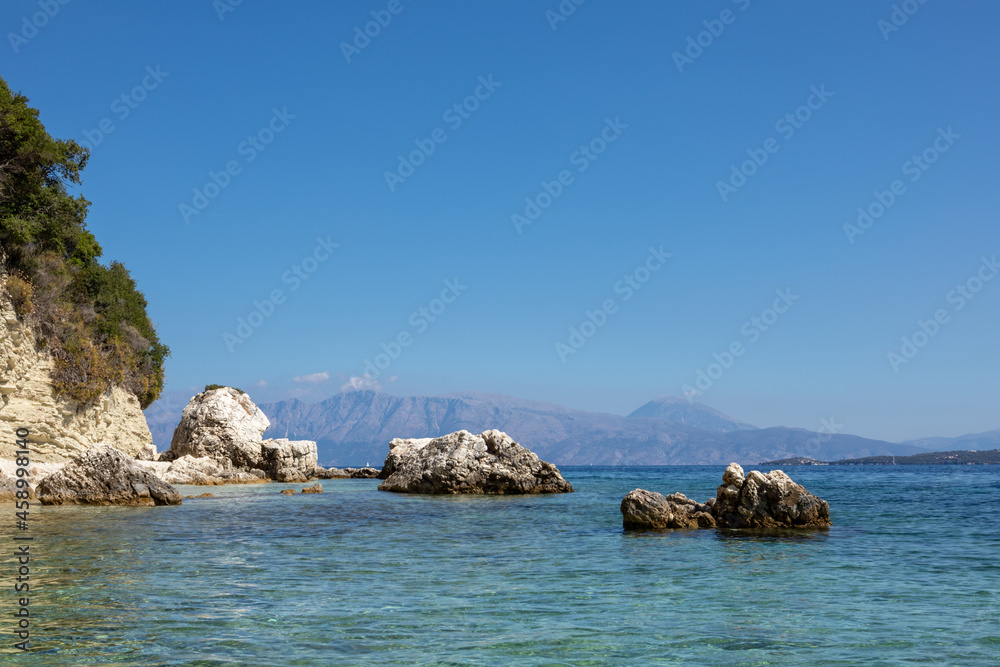 Scenic rocks in blue water on pebble beach of Ionian Sea with distant mountains and bright sky. Nature of Lefkada island in Greece. Summer vacation travel destination