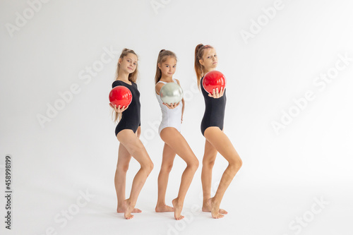 three slim artistic teenager girls in black and white leotards trains and having fun with balls on white background in rhythmic gymnastic exercise, children's professional sports