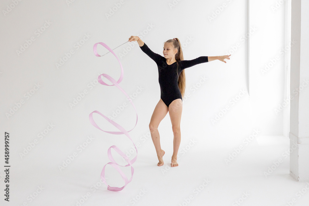 slim artistic teenager girl in black leotard trains on white background with ribbon in her hands in rhythmic gymnastic exercise, children's professional sports